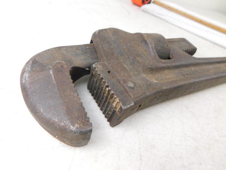 May include: Close-up of a well-worn, rusty pipe wrench. The wrench is made of metal and has a curved jaw and a serrated grip. The tool is designed for gripping and turning pipes and other cylindrical objects.