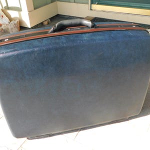 May include: A vintage blue suitcase with a brown trim and a black handle. The suitcase has a rectangular shape and appears to be made of a durable material. The suitcase is closed and ready for travel.