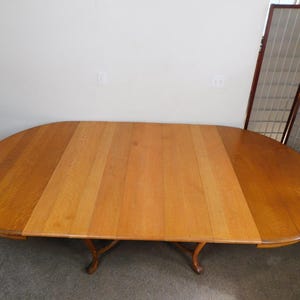 May include: An oval wooden dining table with a light brown finish, featuring a central leaf insert that is lighter in color than the two rounded end sections. The table is supported by curved wooden legs that extend outward from the center base.