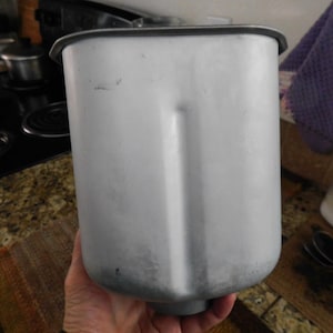 May include: A light gray bread machine pan with a black rim. The pan is cylindrical with a rectangular indentation on one side. The pan is held by a person's hand.