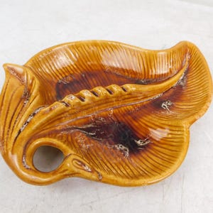 May include: A glossy, glazed ceramic dish shaped like a large leaf with a prominent central vein and a circular handle opening. The surface features a gradient of warm amber, golden yellow, and deep brown tones with a textured, ribbed finish.