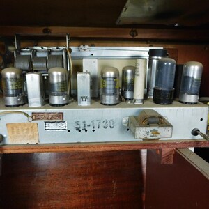 May include: The internal chassis of a vintage radio featuring multiple glass vacuum tubes, a metal tuning capacitor, and a transformer. The metal frame is stamped with the text 51-1730 and includes various electrical components, wiring, and paper labels.