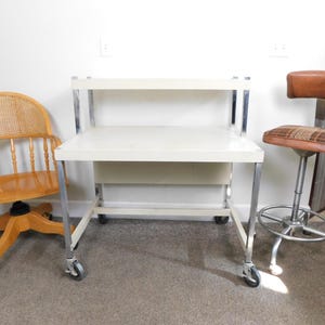 May include: An office scene featuring a light-colored desk on wheels, a wooden swivel chair with a cane back, and a brown and plaid adjustable stool. The desk has a white top and chrome legs.