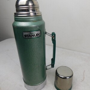 May include: A vintage green Stanley Aladdin thermos with a textured finish, a dark green plastic handle, and a matching dark green screw-on cap. The metal cup lid sits separately beside the thermos, which features a silver Stanley Aladdin label.
