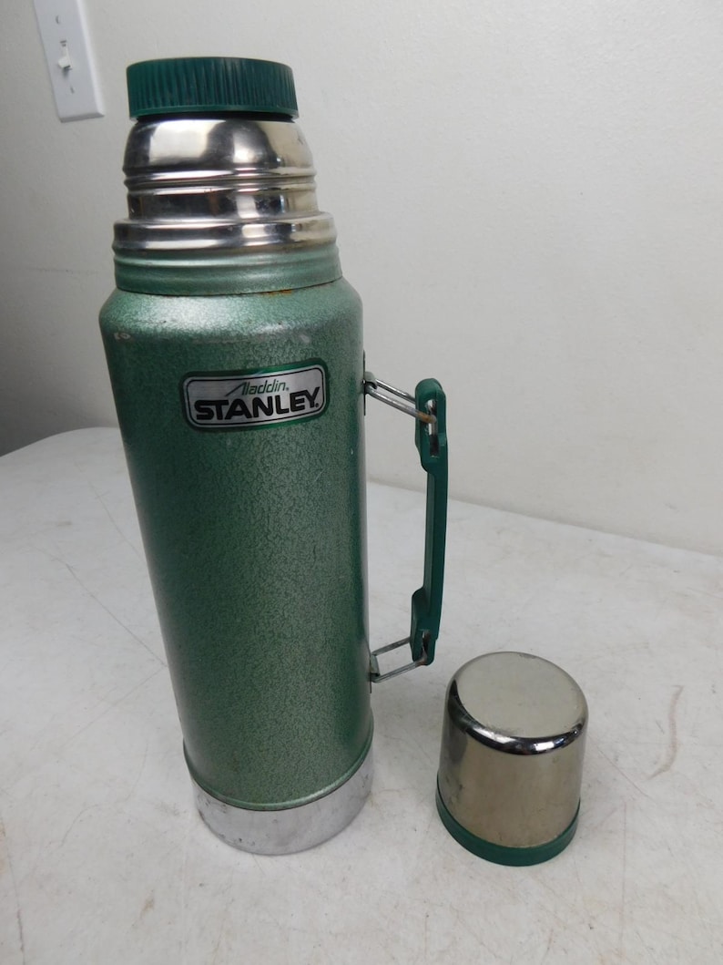 May include: A vintage green Stanley Aladdin thermos with a textured finish, a dark green plastic handle, and a matching dark green screw-on cap. The metal cup lid sits separately beside the thermos, which features a silver Stanley Aladdin label.