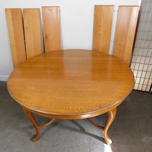 May include: A round wooden table with a light brown finish, featuring curved legs and a matching set of extension leaves. The table is approximately 48 inches in diameter.