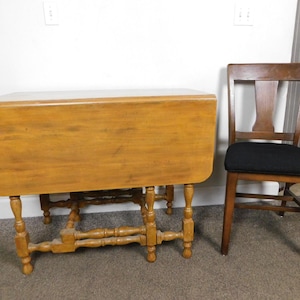 May include: A wooden drop-leaf table with a light brown finish and turned legs. A wooden chair with a black seat cushion is next to the table. The table is closed, and the chair is made of wood.
