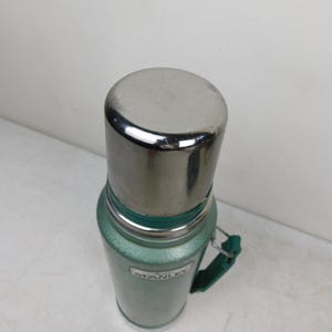 May include: A green Stanley vacuum insulated thermos with a metallic silver cap and a green side handle. The cylindrical body features a textured finish and a white rectangular label with the brand name Stanley printed in black lettering on the front.