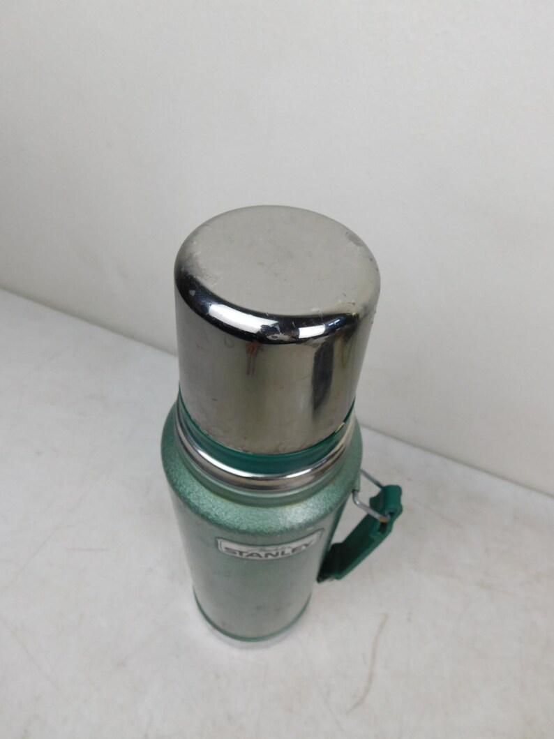 May include: A green Stanley vacuum insulated thermos with a metallic silver cap and a green side handle. The cylindrical body features a textured finish and a white rectangular label with the brand name Stanley printed in black lettering on the front.