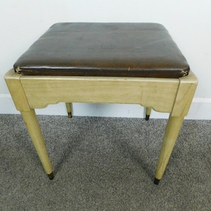 May include: A square stool featuring a dark brown padded seat with visible wear on the edges. The frame is constructed from light-colored wood with a smooth finish, supported by four tapered legs that end in small, dark-colored metal feet.