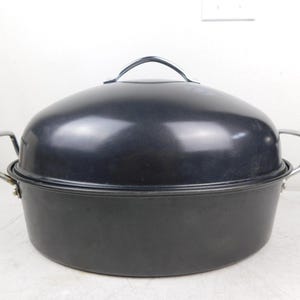 May include: A black oval-shaped roasting pan with a matching lid and silver-colored handles. The lid has a single handle on top. The pan is designed for cooking and baking. The pan is made of metal.