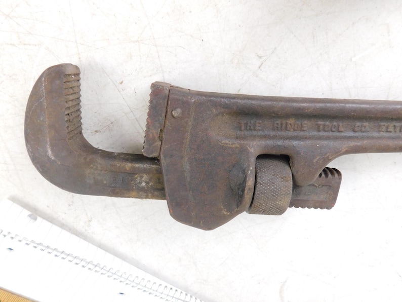 May include: A rusty, vintage pipe wrench with the text "THE RIDGE TOOL CO. ELYRIA" visible on the side. The wrench has a curved jaw and an adjustable jaw, and is likely made of steel.