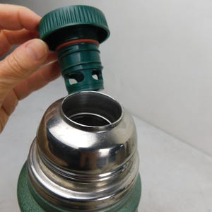 May include: A green vacuum insulated thermos with a metallic silver neck and a removable green plastic stopper. The stopper features a ribbed edge, a red silicone gasket, and a hollow base with circular cutouts for liquid flow control.
