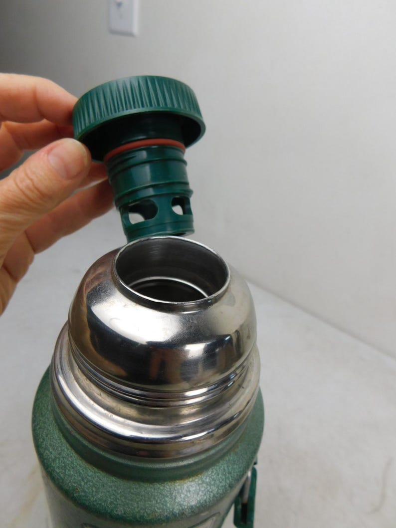 May include: A green vacuum insulated thermos with a metallic silver neck and a removable green plastic stopper. The stopper features a ribbed edge, a red silicone gasket, and a hollow base with circular cutouts for liquid flow control.