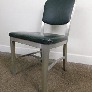 May include: A vintage chair with a dark green padded seat and backrest. The chair has a silver metal frame and legs. The seat and back appear to be made of a textured vinyl material. The chair is designed for seating.