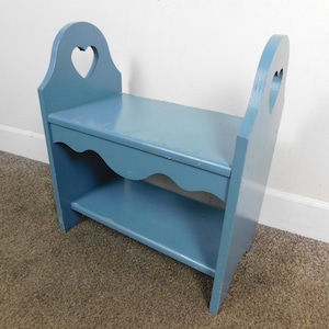 May include: A blue wooden bench with a heart-shaped cutout in the backrest. The bench has a shelf underneath the seat and a scalloped edge. The bench is painted in a solid blue color and is likely for decorative or functional use.