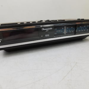 May include: Black Chromatic 251 AM/FM radio alarm clock with a silver trim. The clock face displays FM and AM radio frequency scales, with alarm indicators on the left. The clock has a rectangular shape.