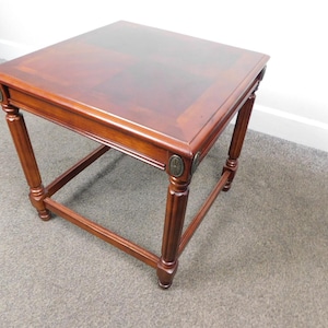 May include: A square wooden side table with a polished mahogany finish. The table features four turned legs connected by a lower support frame and is accented with small, oval-shaped metallic medallions at the top of each leg corner.