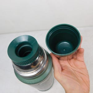 May include: A green vacuum-insulated thermos with a metallic silver neck and a dark green plastic stopper. The thermos features a matching dark green plastic cup lid that is detached and held to the side, revealing the inner threading and opening.
