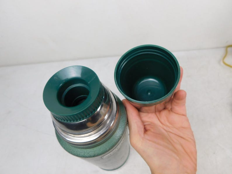May include: A green vacuum-insulated thermos with a metallic silver neck and a dark green plastic stopper. The thermos features a matching dark green plastic cup lid that is detached and held to the side, revealing the inner threading and opening.