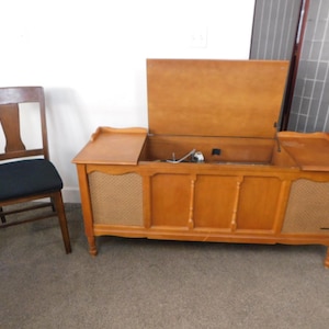 May include: A vintage wooden console stereo with an open top, revealing internal components. The stereo has a light brown finish, decorative panels, and woven speaker covers. A wooden chair with a black seat sits to the left.