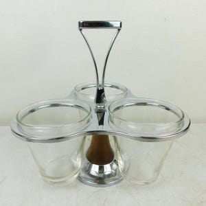 May include: A vintage condiment server with three clear glass cups, a chrome handle, and a wooden base. The cups are held in place by a chrome frame. The handle is a chrome arch. The server is designed to hold condiments.