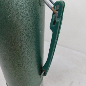 May include: A green vacuum insulated thermos with a textured hammer-tone finish. The cylindrical container features a dark green plastic handle attached with metal hinges. A silver-colored metal base is visible at the bottom of the vessel.