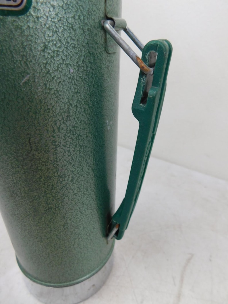 May include: A green vacuum insulated thermos with a textured hammer-tone finish. The cylindrical container features a dark green plastic handle attached with metal hinges. A silver-colored metal base is visible at the bottom of the vessel.