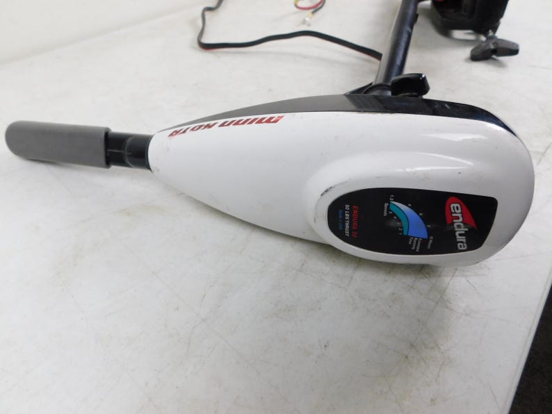 May include: A white and black Minn Kota Endura trolling motor. The motor has a gray handle and a black shaft. The motor has a gauge with blue and green indicators and the brand name "endura" in red.