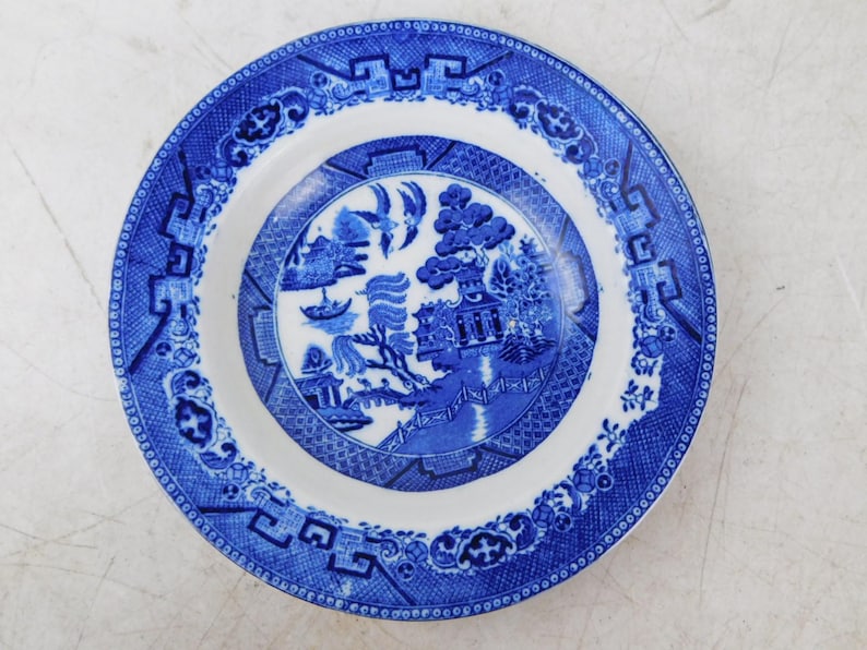 K&ouml;nnte beinhalten: Ein runder, wei&szlig;-blauer Keramikteller mit Willow-Muster. Der Teller zeigt eine detaillierte Landschaftsszene in der Mitte, umgeben von einem blauen Rand mit geometrischen und floralen Motiven. Ein klassisches St&uuml;ck Geschirr.