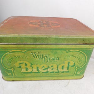 May include: A rectangular metal bread box with a green finish and a rust-colored lid. The front features the text Wheat Heart Brand Bread, Oven-Baked, and Enriched. The lid has a central decorative emblem. The container has a vintage appearance.