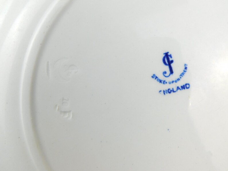 K&ouml;nnte beinhalten: Wei&szlig;er Keramikteller mit gew&ouml;lbtem Rand. Der Teller hat blauen Text mit der Aufschrift "Stoke-upon-Trent" und "England". Es gibt auch ein blaues Logo und andere Markierungen.