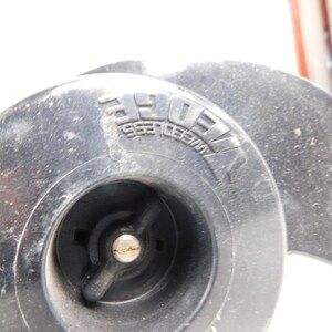 May include: Close-up of a black, circular object with the embossed text "WEEDLESS" visible. The object has a central hub with a metal fitting and a propeller-like design. The overall appearance suggests a mechanical component.