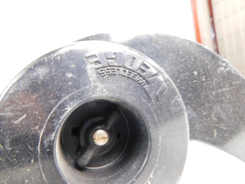 May include: Close-up of a black, circular object with the embossed text "WEEDLESS" visible. The object has a central hub with a metal fitting and a propeller-like design. The overall appearance suggests a mechanical component.