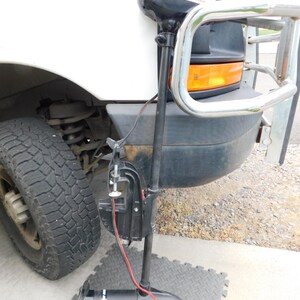 May include: A black trolling motor with red wires is mounted on the front of a white truck. The motor is positioned on a gray, diamond-patterned mat. The truck has a chrome bumper guard and a black front end.