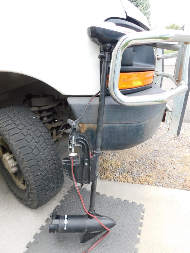 May include: A black trolling motor with red wires is mounted on the front of a white truck. The motor is positioned on a gray, diamond-patterned mat. The truck has a chrome bumper guard and a black front end.