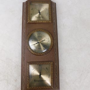 May include: A rectangular wall-mounted weather station featuring a brown wood-tone frame with decorative edges. It contains three gold-toned circular and square gauges: a top humidity meter, a central barometer, and a bottom temperature gauge.