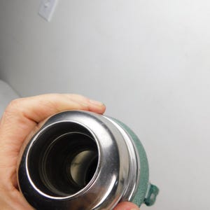 May include: A top-down view of a cylindrical metal thermos with a polished silver-colored rim and a textured sage green exterior finish. The opening reveals a dark interior cavity, and a small green plastic handle is visible on the side.