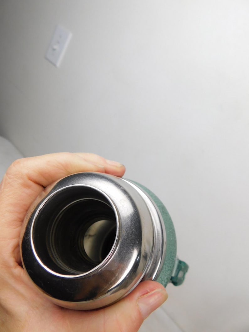 May include: A top-down view of a cylindrical metal thermos with a polished silver-colored rim and a textured sage green exterior finish. The opening reveals a dark interior cavity, and a small green plastic handle is visible on the side.