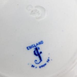 K&ouml;nnte beinhalten: Nahaufnahme eines wei&szlig;en Keramiktellers mit blauem Stempel. Der Stempel lautet "ENGLAND" &uuml;ber einem stilisierten "J" und anderem Text. Der Teller hat ein schlichtes, elegantes Design, das sich zum Servieren von Speisen eignet.