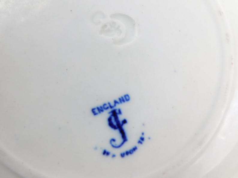 K&ouml;nnte beinhalten: Nahaufnahme eines wei&szlig;en Keramiktellers mit blauem Stempel. Der Stempel lautet "ENGLAND" &uuml;ber einem stilisierten "J" und anderem Text. Der Teller hat ein schlichtes, elegantes Design, das sich zum Servieren von Speisen eignet.