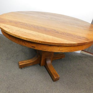 May include: A round wooden table with a light brown finish. The table has a pedestal base with four legs. The tabletop is approximately 48 inches in diameter. The table is made of solid wood and has a classic design.