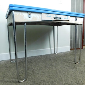 May include: A vintage metal table featuring a bright blue rectangular top and a cream-colored apron with a single central drawer. The table is supported by four slender, U-shaped chrome legs that provide a stable base for the structure.