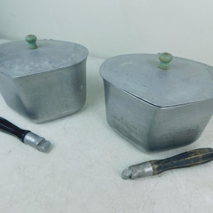 May include: Two silver-colored metal containers with matching lids, each featuring a small, light green knob. Two detachable black handles with metallic ends are positioned in front of the containers, which have a rounded, trapezoidal shape.