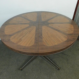 May include: A round dining table featuring a two-tone wood veneer top arranged in a radial pattern resembling citrus segments. The table is supported by a central metal base with four splayed legs that extend outward from the center point.