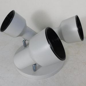 May include: A white, three-light spotlight fixture with black interior cones. The lights are mounted on a circular base with silver-colored supports. The fixture is designed to direct light.