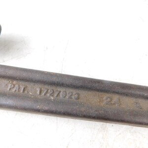May include: Close-up of a vintage pipe wrench. The wrench is made of dark metal with visible wear and tear. The text "PAT. 1727023" is stamped on the side, indicating a patent number. The tool is designed for gripping and turning pipes.
