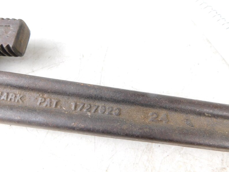 May include: Close-up of a vintage pipe wrench. The wrench is made of dark metal with visible wear and tear. The text "PAT. 1727023" is stamped on the side, indicating a patent number. The tool is designed for gripping and turning pipes.