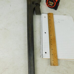 May include: A large, dark gray pipe wrench, a black and orange tape measure, a wooden ruler, and a spiral notebook with lined paper. The tape measure reads "25". The ruler is marked in inches.