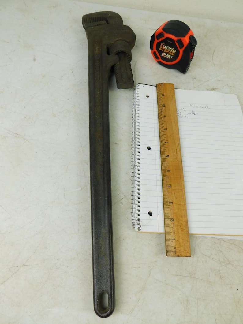 May include: A large, dark gray pipe wrench, a black and orange tape measure, a wooden ruler, and a spiral notebook with lined paper. The tape measure reads "25". The ruler is marked in inches.